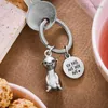 A close-up shot of a silver meerkat keychain with a round charm attached, resting on a brown textured surface next to a key and a partial view of a book and a cup of tea. Produktbild-Vorschau