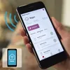 A person is holding a smartphone displaying a smart home app interface. The app shows weather information and controls for various devices like music, ventilation, and lighting. Produktbild-Vorschau