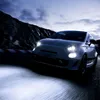 A white car with its headlights on, driving on a road at dusk. Produktbild-Vorschau