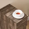 A close-up shot of a white teacup and saucer filled with tea, placed on the top surface of a dark brown wooden nightstand. Anteprima dell’immagine prodotto