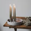 A round, light gray candle holder with four lit taper candles sits on a rustic wooden table next to a dried floral arrangement. Produktbild-Vorschau