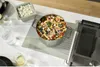 A pasta salad is presented in a stainless steel pot on a kitchen counter next to a container of feta cheese and a glass of water with lemon. Voir les images du produit