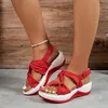 Person trägt rote Keilabsatz-Sandalen mit geknoteten Riemen und weißen Sohlen, steht auf grauem Boden Náhled obrázku produktu