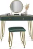 A dark green vanity table with gold legs and a round mirror with LED lighting, along with a matching stool, is displayed against a white background. Decorative items like makeup and a vase with dried flowers are placed on the table. Náhľad obrázku produktu