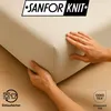 Hands are shown adjusting the corner of a beige fitted sheet, demonstrating its fit and material. The background is a wooden surface. Produktbild-Vorschau
