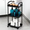 A black rolling cart with three shelves, containing toiletries and toilet paper, placed on a tiled floor next to a white cabinet. Anteprima dell’immagine prodotto