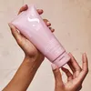 Hands holding a pink tube of conditioner against a light, textured background. Produktbild-Vorschau