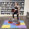 A woman is exercising on a step platform in a room with bookshelves in the background. Voir les images du produit