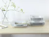 A set of white square plates and bowls are neatly stacked on a wooden table, with a vase of greenery in the background. Náhľad obrázku produktu