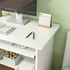 A close-up view of a white computer desk with a Dell monitor, a calendar, and pens on top. Shelving below the desk holds a decorative box filled with round objects. Produktbild-Vorschau