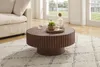 A round coffee table with a fluted base is placed on a rug in a living room setting, next to a sofa. A carafe of water and a small potted plant are on the table. Produktbild-Vorschau