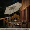 A patio umbrella with solar LED lights is set up on a wooden deck at night, next to a wooden house with red doors. There are chairs and a table on the deck, and a small animal is visible. Náhled obrázku produktu