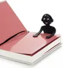 A black bookmark in the shape of a person with headphones is placed on an open book with pink pages. Voir les images du produit