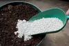 A hand is adding white granular material from a green scoop into a pot of dark soil. Produktbild-Vorschau