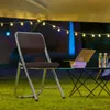 A folding chair and table with a laptop and water bottles set up on grass under string lights. Produktbild-Vorschau