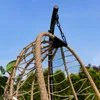 A close-up view of the top of a hanging chair, showing the woven material and the metal stand it hangs from, against a blue sky and green trees. Produktbild-Vorschau