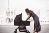 A woman in a dress is bending over a black baby stroller in a public square with a large building in the background. Produktbild-Vorschau