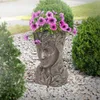 A bronze-colored planter shaped like a woman's head, filled with purple petunias, sits on a bed of gravel surrounded by greenery. Náhled obrázku produktu