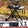 A portable gas grill is set up on a folding table outdoors, with autumn leaves on the ground. Produktbild-Vorschau