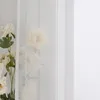 White flowers are visible behind a sheer white curtain, with a white wall on the right. Produktbild-Vorschau