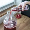 A hand is pouring red liquid from a bottle into a large glass carboy. Produktbild-Vorschau