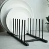 White plates are stacked on a black metal plate rack on a light gray surface. Produktbild-Vorschau
