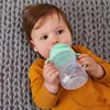 A baby lying on a gray knitted blanket, drinking from a green sippy cup. Produktbild-Vorschau