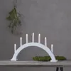 A white arched candle holder with seven lit LED candles sits on a wooden table. A sprig of greenery hangs from the ceiling in the background. Produktbild-Vorschau