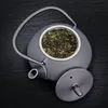 A top-down view of a black cast iron teapot filled with green tea leaves, with its lid placed beside it on a dark, textured surface. Podgląd zdjęcia produktu