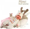 A baby doll dressed in a deer-themed outfit, lying on a pink blanket next to a deer-shaped pillow. Podgląd zdjęcia produktu