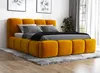 A yellow upholstered bed with gray bedding is shown in a room with artwork on the wall and a plant in the background.