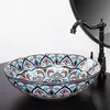 A decorative ceramic sink with a faucet and a mirror in the background, placed on a black countertop. Náhled obrázku produktu