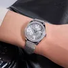 A close-up shot of a woman's wrist wearing a silver Guess watch with a diamond-encrusted bezel and a patterned dial. The background is dark and out of focus, suggesting a lifestyle setting. Náhled obrázku produktu
