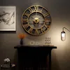 A large, vintage-style wall clock with Roman numerals is displayed on a dark gray wall above a black mantelpiece. Decorative items, including a vase with dried flowers and small sculptures, are arranged on the mantelpiece. A modern lamp with a white shade  Produktbild-Vorschau