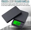 A black digital scale with a green display showing '0.00 g'. The scale is shown with its lid open, revealing the weighing platform. The background is a light gray and white abstract design. Náhľad obrázku produktu