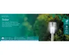A solar garden stake light is shown in a natural outdoor setting with greenery in the background. Produktbild-Vorschau