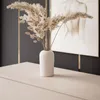 A vase with dried flowers sits on a light-colored surface in front of a dark wall with gold accents and a framed artwork. Produktbild-Vorschau