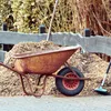 A rusty wheelbarrow filled with wood chips sits next to a pile of wood chips and a broom. Produktbild-Vorschau