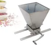 A stainless steel manual grain mill with a hopper and a crank handle, placed on a white background with some scattered beans and bowls of beans on the left side. Podgląd zdjęcia produktu