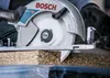 A close-up shot of a circular saw blade cutting through a piece of particle board. Voir les images du produit