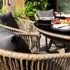 A close-up view of a wicker outdoor dining set with dark gray cushions, set on a patio with greenery in the background. Produktbild-Vorschau