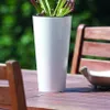 A white planter with a plant sits on a wooden table next to a wooden chair. Anteprima dell’immagine prodotto