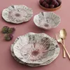 A set of decorative plates and bowls with a floral-like pattern, along with grapes and flowers, arranged on a pink tablecloth. Produktbild-Vorschau