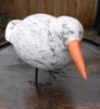 A decorative wooden bird sculpture with an orange beak and black wire legs, placed on a wooden surface. Produktbild-Vorschau