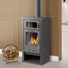 A dark gray wood-burning stove with a glass door showing flames, placed in a room with tan walls and white baseboards. A basket of firewood is to the left. Produktbild-Vorschau