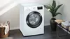 A white front-loading washing machine is shown in a clean, modern laundry room setting, with a basket of clothes and a plant nearby. Produktbild-Vorschau