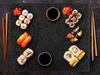A top-down view of various sushi dishes arranged on a dark, textured surface, with chopsticks and soy sauce dishes. Náhľad obrázku produktu
