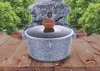A granite-style pot with wooden handles and lid knob, set against a backdrop of trees and a rock with text. Náhľad obrázku produktu