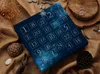 An advent calendar with numbered squares is placed on a burlap fabric background, surrounded by natural elements like stones, wood, and dried plants. Anteprima dell’immagine prodotto