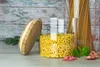 A glass jar filled with elbow macaroni is placed on a wooden surface, with its bamboo lid resting beside it. Other types of pasta and a sprig of basil are also visible. Produktbild-Vorschau